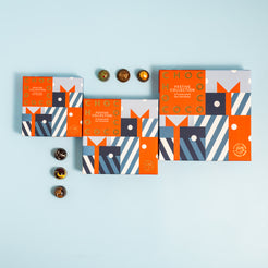 Three decorative chocolate gift boxes arranged on a light blue background, with several round chocolates placed beside them. The boxes feature a geometric pattern in red, navy, blue, white, and beige, with the words 'FESTIVE COLLECTION' visible on the front.