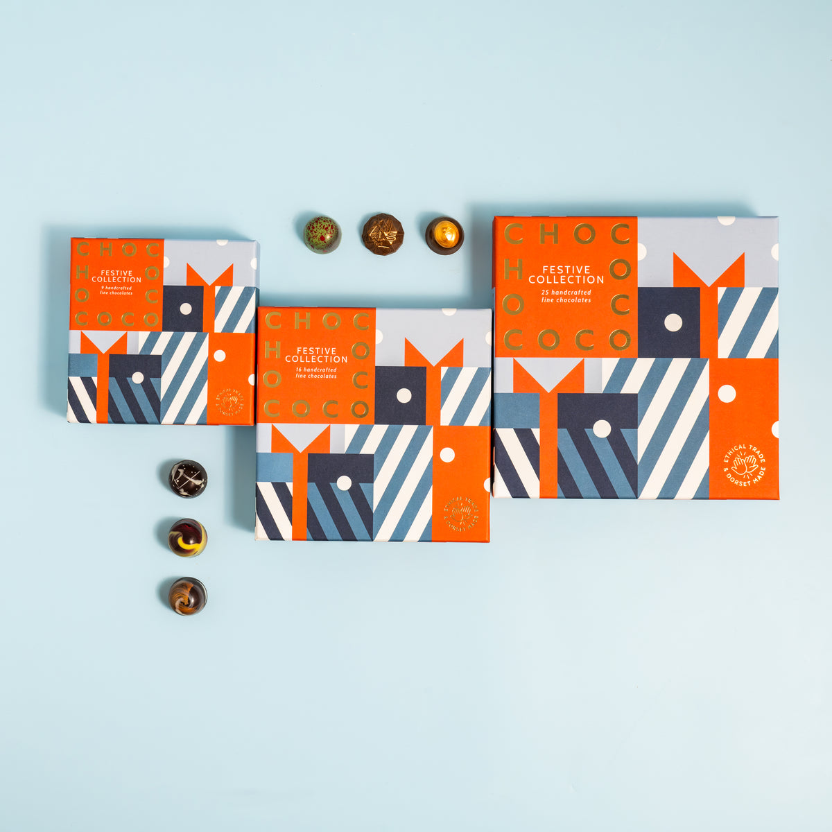 Three decorative chocolate gift boxes arranged on a light blue background, with several round chocolates placed beside them. The boxes feature a geometric pattern in red, navy, blue, white, and beige, with the words 'FESTIVE COLLECTION' visible on the front.