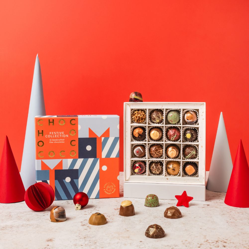A festive chocolate collection gift set displayed on a white surface against a solid red-orange background. The set includes a colorful patterned box labeled 'FESTIVE COLLECTION', an open white box filled with assorted chocolates, and several loose chocolates scattered in front.