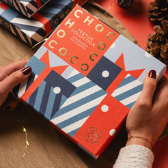 A person holds a square gift box for the 'Chococo Festive Collection' with a colorful geometric holiday pattern. The box features red, navy blue, light blue, and white shapes with gold foil lettering.