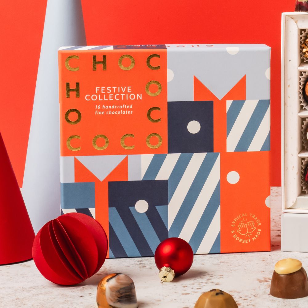 A boxed chocolate collection called 'CHOCOCO FESTIVE COLLECTION' sits on a light surface with red and gold Christmas props. The box has a colorful geometric pattern in red, navy, white, and blue, with gold lettering.