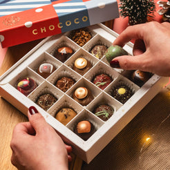 An open box of assorted artisan chocolates on a wooden table, with a hand placing a green decorated chocolate into the tray. The red and blue box lid in the background shows the brand name Chococo