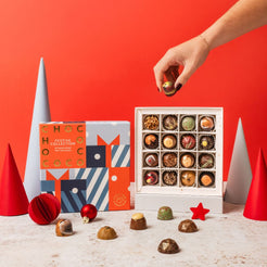 A hand reaches for a chocolate from an open white box of assorted chocolates, with a Chococo branded gift box beside it and several chocolates scattered on a light surface against a bright red background.