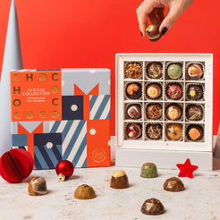 A boxed assortment of chocolates displayed on a light surface against a bright red background, with a hand holding one chocolate above an open white tray. The packaging includes a colorful box labeled 'CHOCOCO FESTIVE COLLECTION' and several loose chocolates in front.
